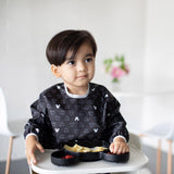 A dark-haired child in a high chair, wearing a Bumkins Sleeved Bib: Mickey Mouse Icon Black + White, eats from a black Mickey Mouse plate.