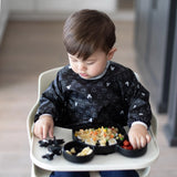 A toddler in a high chair in a Bumkins Sleeved Bib: Mickey Mouse Icon Black + White eats from a divided plate filled with rice and fruit.