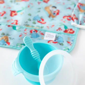 Close-up of a blue baby bowl and spoon on a white background, with Bumkins Sleeved Bib: Ariel blurred behind.
