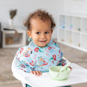 A curly-haired toddler in a high chair wears a Bumkins Sleeved Bib: Ariel with a green bowl and spoon on the tray.
