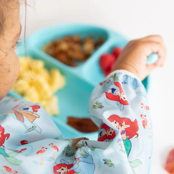 Close-up of a child enjoying snacks from a blue plate with a fork, wearing Bumkins Sleeved Bib: Ariel.