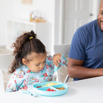 A child in a Bumkins Sleeved Bib: Ariel eats from a blue plate with a fork, sitting beside a smiling man at a white table.