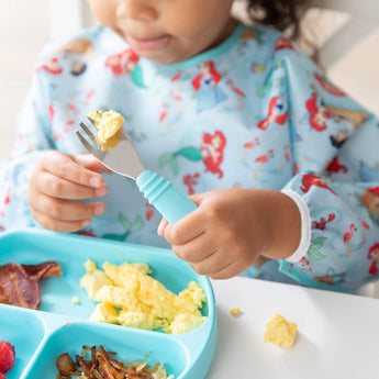 A child in a Bumkins Sleeved Bib: Ariel holds a fork with scrambled eggs by a blue sectioned breakfast plate at a white table.