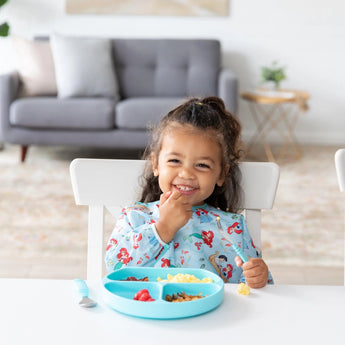 A child wearing Bumkins Sleeved Bib: Ariel smiles while eating from a blue sectioned plate at a white table; a gray couch in the background.