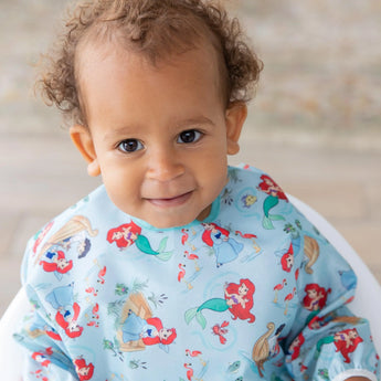 A toddler smiles at the camera, wearing a colorful Bumkins Sleeved Bib: Ariel with cartoon mermaids.