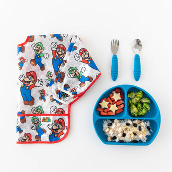 Bumkins Sleeved Bib: Super Mario™ Classic lays folded next to a blue plate with fruit, broccoli, and rice, and utensils on a white background.