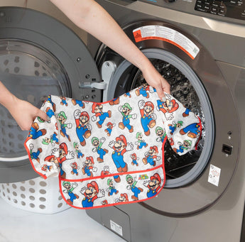 A person holds Bumkins Sleeved Bib: Super Mario™ Classic in front of an open front-loading washing machine.