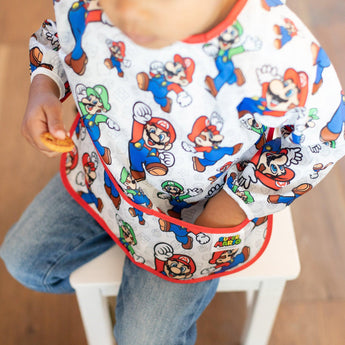 A child in a Bumkins Sleeved Bib: Super Mario™ Classic sits on a white chair with their hand in the pocket.