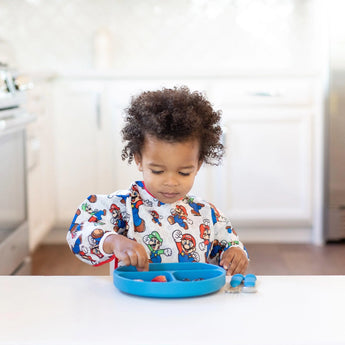 A toddler in a Bumkins Sleeved Bib: Super Mario™ Classic enjoys food from a blue plate at a white table in a kitchen.
