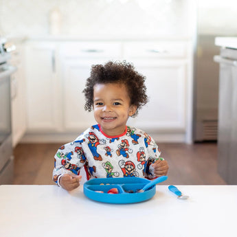 A toddler smiles at a white table, wearing a Bumkins Sleeved Bib: Super Mario™ Classic and eating from a blue plate in a bright kitchen.