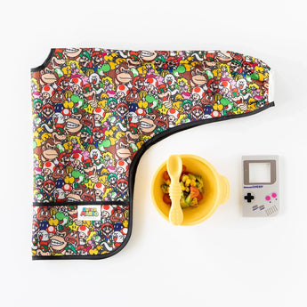 Bumkins Sleeved Bib: Super Mario™ Mashup lays folded by a yellow bowl with spoon, and Nintendo Game Boy teether on a white background.