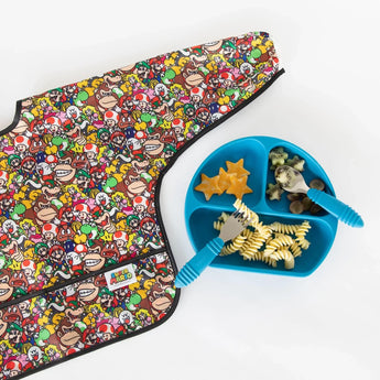 Bumkins Sleeved Bib: Super Mario™ Mashup lays flat beside a blue divided plate with pasta, cheese, fruit, and utensils on a white background.