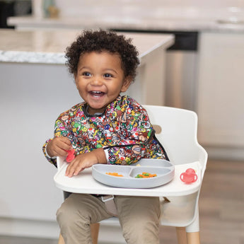 A happy toddler in a Bumkins Sleeved Bib: Super Mario™ Mashup holds a red toddler spoon, sitting in a high chair with food on a gray plate.