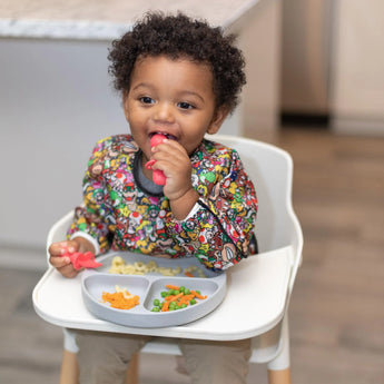 A toddler in a Bumkins Sleeved Bib: Super Mario™ Mashup eats happily from a gray plate in a high chair, holding a red toddler spoon.