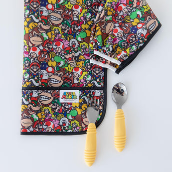 Bumkins Sleeved Bib: Super Mario™ Mashup lays folded near a yellow-handled spoon & fork on a white background.