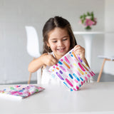 A girl in a white shirt smiles, holding a Bumkins Reusable Snack Bag, Large 2-Pack: Watercolor & Brush Strokes as the other sits on a table.