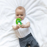 A baby in white outfit on a white blanket holds a Bumkins Silicone Teether: Dinosaur.