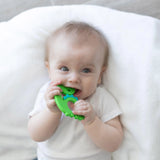 A baby chews on a Bumkins Silicone Teether: Dinosaur, lying on a white blanket.