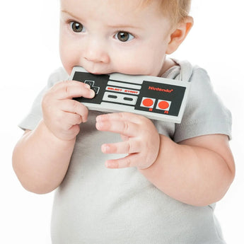 A baby in a gray shirt chews on a Bumkins Silicone Teether: NES™ Controller on a white background.