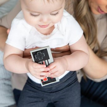A baby holding a Bumkins Silicone Teether: NES™ Controller sits on a woman's lap.
