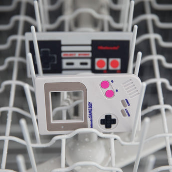 Bumkins Silicone Teether: NES™ Controller  sits behind another teether in the top rack of an open dishwasher.