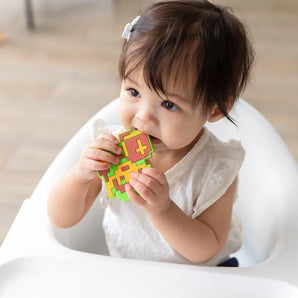 A toddler in a high chair, wearing a white shirt, chews a Bumkins Silicone Teether: Zelda™ Link.