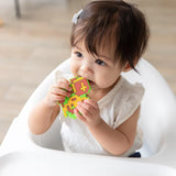 A toddler in a high chair, wearing a white shirt, chews a Bumkins Silicone Teether: Zelda™ Link.