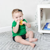 A baby sits on a gray carpet, smiling and chewing a Bumkins Silicone Teether: Zelda™ Link near a white shelf.