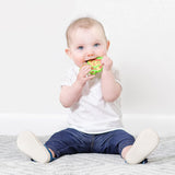 A baby on a carpet in white and blue clothes chews a Bumkins Silicone Teether: Zelda™ Link.