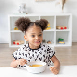A young girl with curly hair wears a Bumkins Junior Bib: Hearts and eats soup from a white bowl with a spoon at a white table.