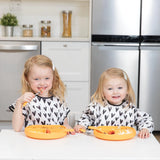 Two kids wearing matching print bibs sit at a white table with orange plates in a kitchen; one wears a Bumkins Junior Bib: Hearts.
