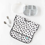 Bumkins Junior Bib: Hearts lays next to a gray plate, transparent lid, and gray utensils on a white background.