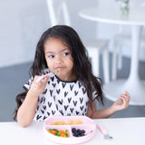 A young girl with long hair wears a Bumkins Junior Bib: Hearts as she eats noodles and veggies from a pink plate, looking thoughtful.