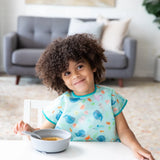 A curly-haired child smiles, wearing a Bumkins Junior Bib: Ocean Life, with a gray bowl and spoon on a white table.