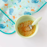 A green bowl of soup with a green spoon sit next to Bumkins Junior Bib: Ocean Life on a white background.