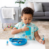 A child in a Bumkins Junior Bib: Ocean Life sips from a blue cup and holds a blue fork at a table with a blue divided plate.