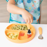 A child in a Bumkins Junior Bib: Ocean Life eating pasta, avocado, and fruit from an orange sectioned plate using an orange fork.