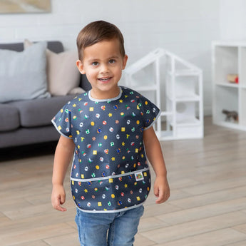In a bright living room, a smiling child wears a Bumkins Junior Bib: Super Mario™ Lineup .