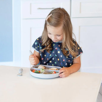 A young girl in a Bumkins Junior Bib: Super Mario™ Lineup sits at a table, eating with utensils from a plate of various foods.
