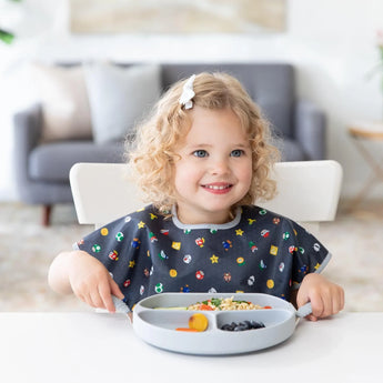 A curly-haired child in a Bumkins Junior Bib: Super Mario™ Lineup sits happily at a white table holding utensils near a gray plate of food.