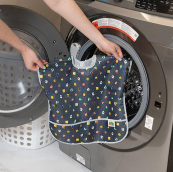 A person holds Bumkins Junior Bib: Super Mario™ Lineup in front of an open front-loading washing machine.
