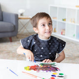 A child painting with watercolors at a white table, wearing a Bumkins Short-Sleeved Smock: Mickey Mouse Icon B+W in a cozy room.