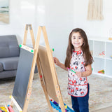 A child painting at an easel in a living room, wearing a Bumkins Short-Sleeved Smock: Minnie Mouse Classic and holding a paintbrush.