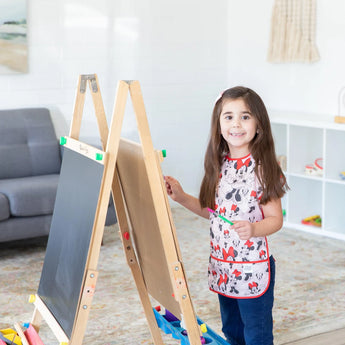 A child painting at an easel in a living room, wearing a Bumkins Short-Sleeved Smock: Minnie Mouse Classic and holding a paintbrush.