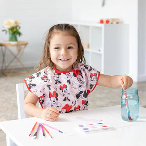 A child smiles while painting in a Bumkins Short-Sleeved Smock: Minnie Mouse Classic, with art supplies scattered on the white table.