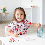 A child smiles while painting in a Bumkins Short-Sleeved Smock: Minnie Mouse Classic, with art supplies scattered on the white table.