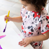 A child painting with a purple brush at a white table, wearing a Bumkins Short-Sleeved Smock: Minnie Mouse Classic.