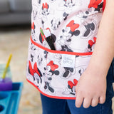 Close-up of a child wearing a Bumkins Short-Sleeved Smock: Minnie Mouse Classic next to an easel, with paintbrushes in the pocket.