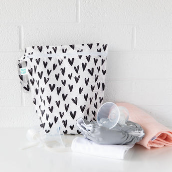 Bumkins Wet Bag: Hearts sits on a white table against a white brick wall, with cloth diapers and breast pump parts in front.