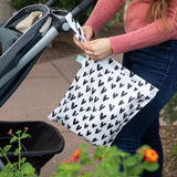 A person attaches a Bumkins Wet Bag: Hearts to a stroller using the strap, while vibrant flowers bloom in the foreground.